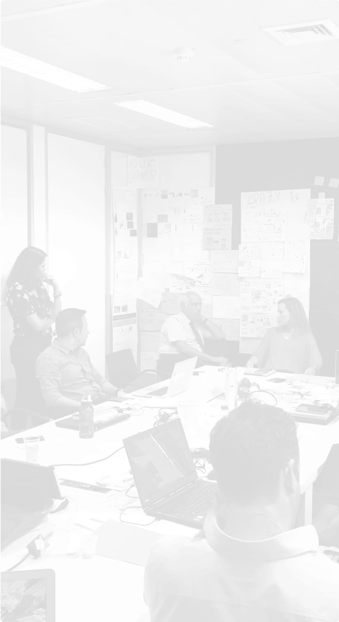 Group of people in a meeting room participating in a brainstorming session. The room is filled with post-it notes and annotations on the walls, and several people are seated around a table with laptops and papers.