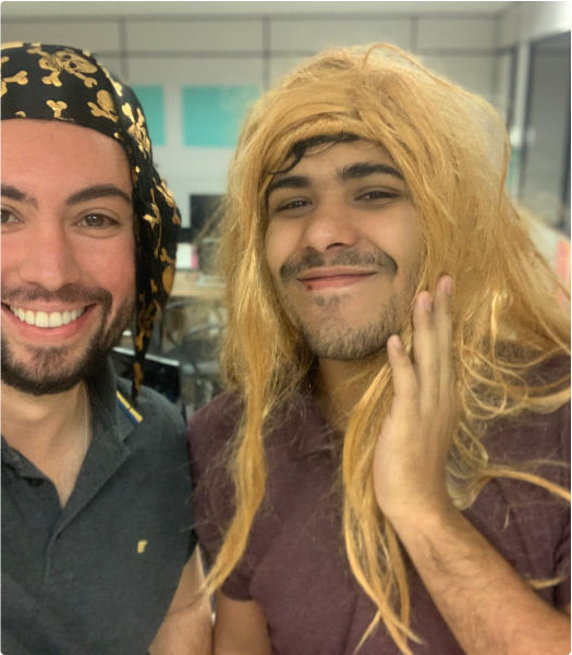 Colleagues having fun with costumes in an office. Two people smiling at the camera, one wearing a black bandana with a gold pattern and the other a blonde wig. Both are in an indoor setting, likely an office.