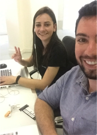 Two colleagues working together and smiling at the camera. Two people sitting at a work desk, smiling at the camera. The person on the left is wearing earphones and making a peace sign with their fingers, while the person on the right is smiling broadly.