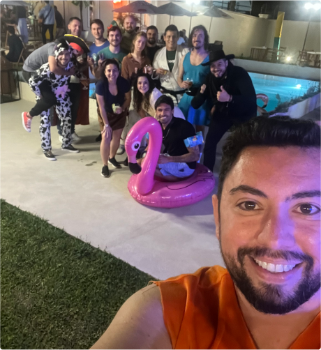 People at a costume party near the pool. A group of people outdoors at night, some wearing costumes. They are posing for a photo near a pool. One person in the front is taking the selfie, while others in the background pose with a pink flamingo float.