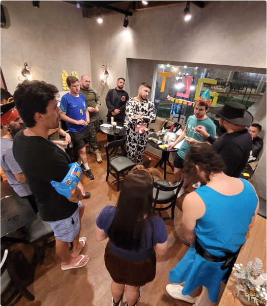 People at a costume social gathering. A group of people in a room, some wearing costumes. They are gathered in a circle, talking and interacting. The room is well-lit and has a large window in the background.