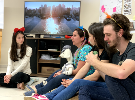 Relaxed brainstorming session with fun accessories. Group of four people sitting on the floor in a room, some wearing fun accessories on their heads. In the background, a TV displays an image of a city at sunset.