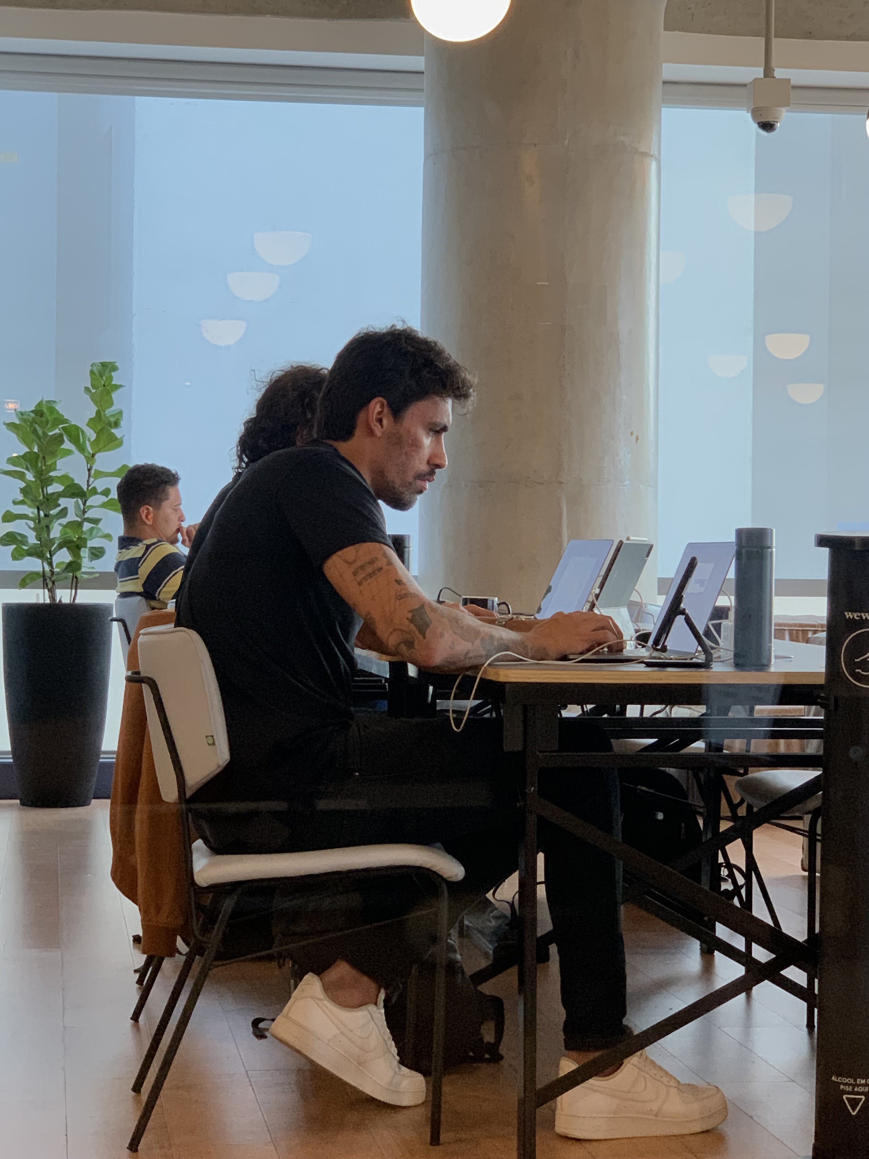Man working in a shared workspace environment. Man focused on working on a laptop at a shared workspace table. There are other people working in the background in a modern environment with large windows.