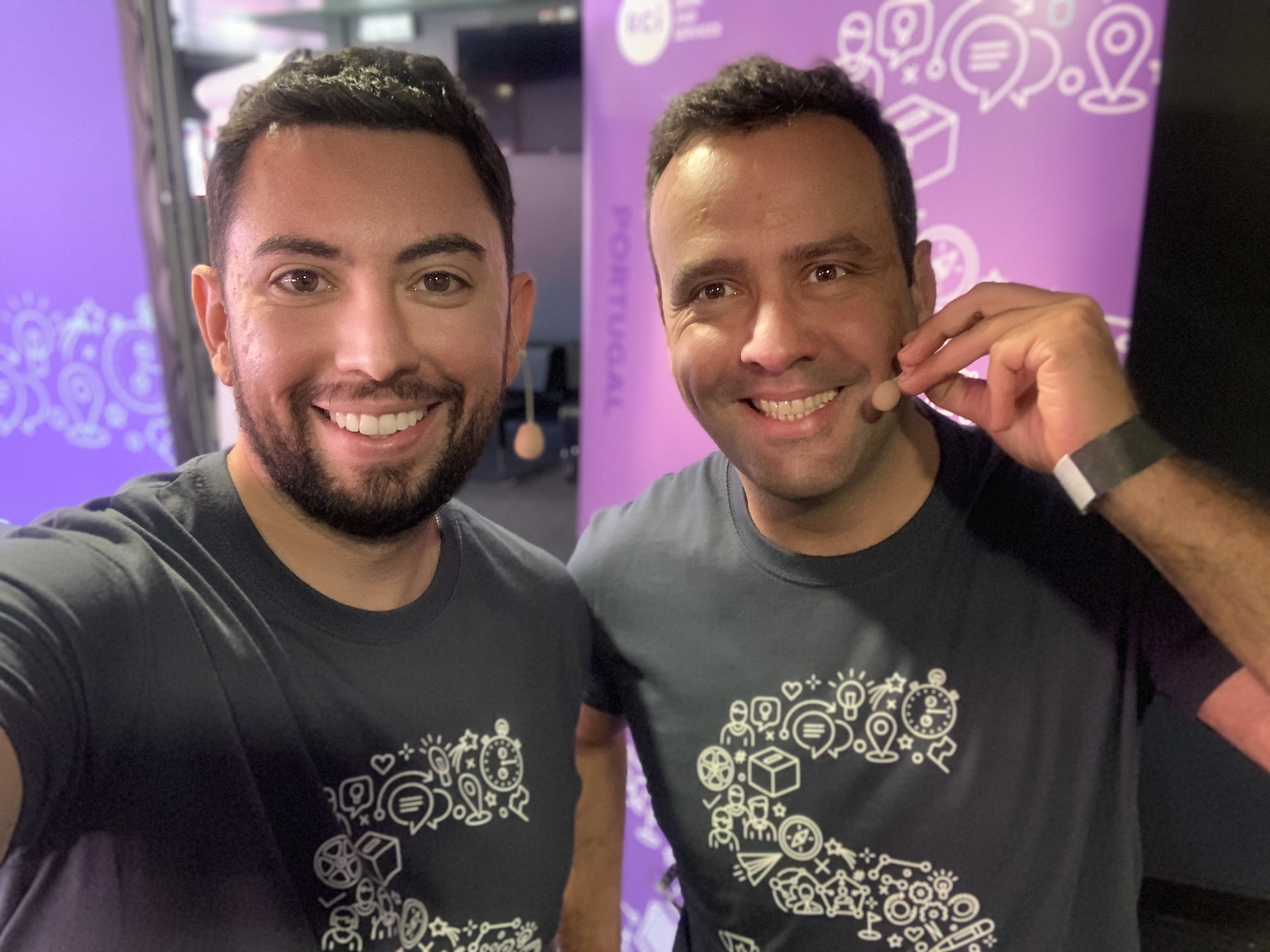 Two men smiling and taking a selfie at an event. Two men smiling and taking a selfie. Both are wearing black t-shirts with a graphic design, and one is wearing a headset microphone. The background is decorated with purple graphics.