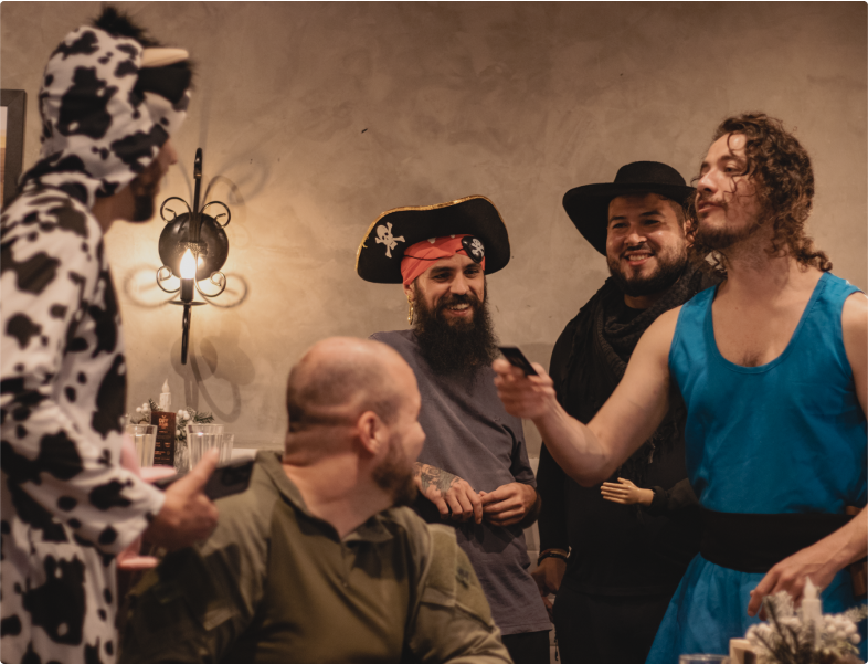 Group of people at a costume party, interacting and having fun. Five people in an indoor setting, all dressed in costumes. One person is in a cow costume, another as a pirate, another with a cowboy hat, and another in a blue costume. They are smiling and talking, creating a cheerful and relaxed atmosphere.