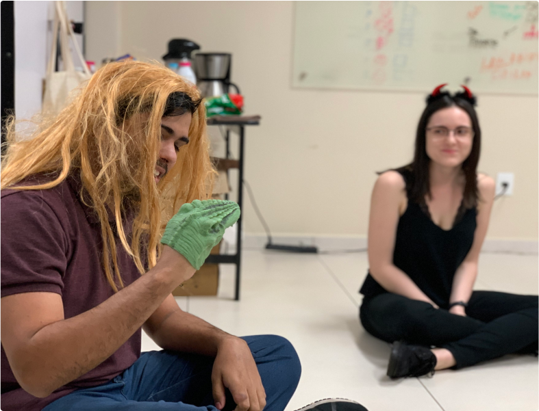 Two people sitting on the floor in an indoor setting. The person on the left is wearing a blonde wig and a green glove, while the person on the right is wearing a headband with red horns. Both are smiling and seem to be having fun. People having fun with costumes indoors.