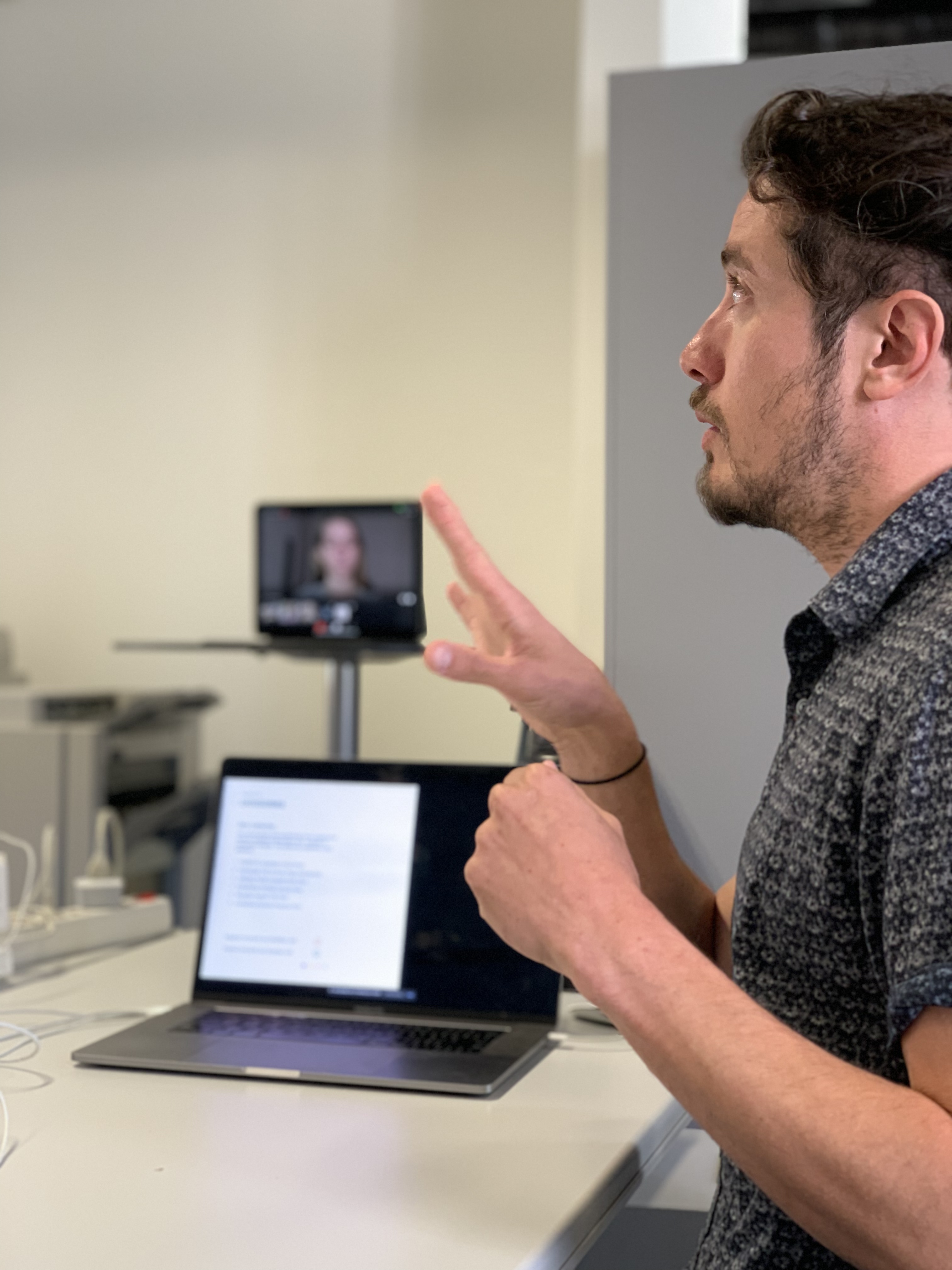 Presentation with video call in an office environment. Man gesturing while giving a presentation, with an open laptop in front of him and a person on a video call visible in the background on the screen.