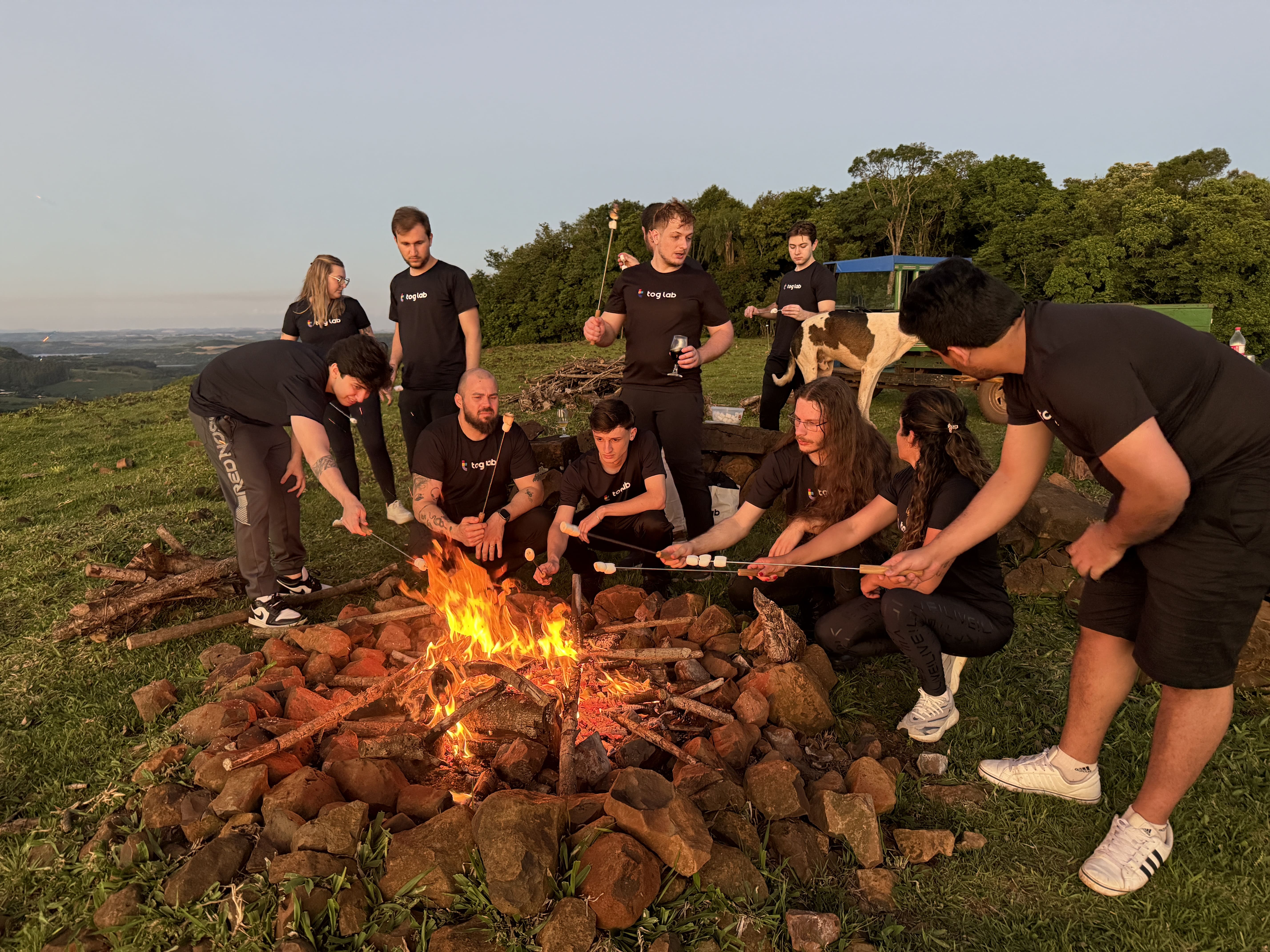 Várias pessoas assando marshmallows em um fogo de acampamento à noite. As mãos seguram espetos com marshmallows sobre as chamas, criando um clima acolhedor e amigável.