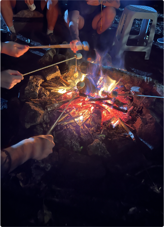 Roasting marshmallows over a campfire at night. Several people roasting marshmallows over a campfire at night. Hands are holding skewers with marshmallows over the flames, creating a cozy and friendly atmosphere.