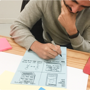 Person creating a wireframe manually in a collaborative work environment. Person drawing a wireframe on a blue sheet of paper at a table. Around the table, there are colorful post-it notes and other stationery materials.