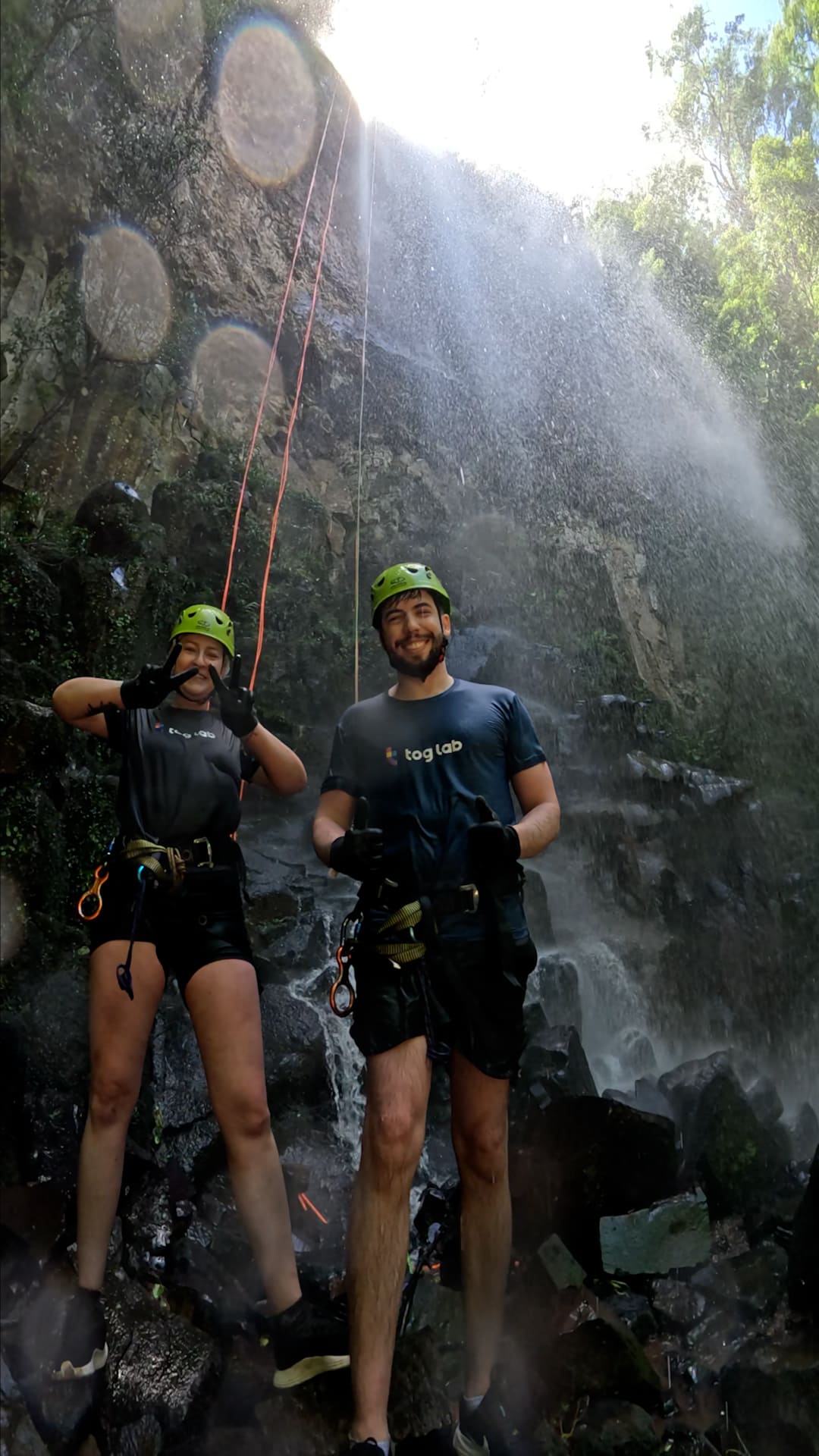 Duas pessoas com capacetes e equipamentos de rapel posam sorrindo sob uma cachoeira em um ambiente natural.