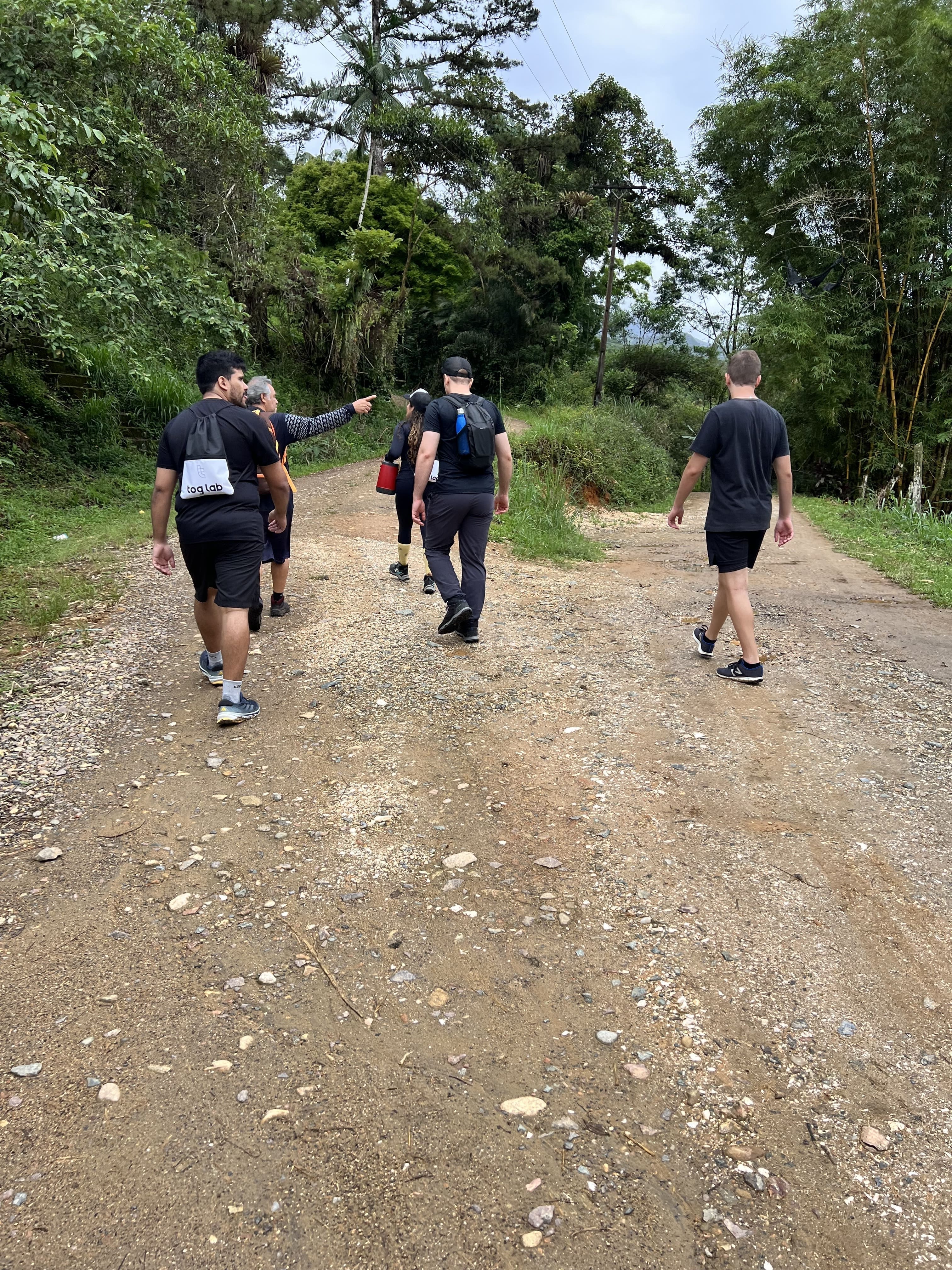 Grupo de membros da equipe caminhando por uma trilha de terra cercada por árvores e vegetação durante uma atividade ao ar livre.