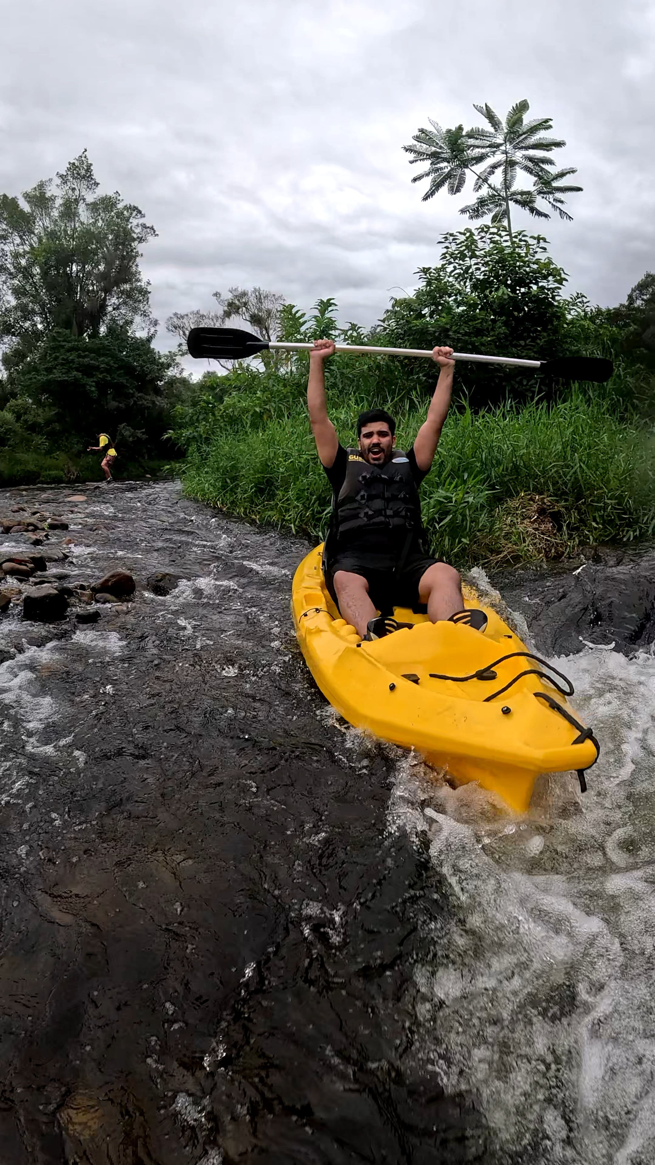 Membro da equipe usando colete salva-vidas descendo de caiaque por um rio raso e rochoso com o remo levantado, cercado por vegetação verde.