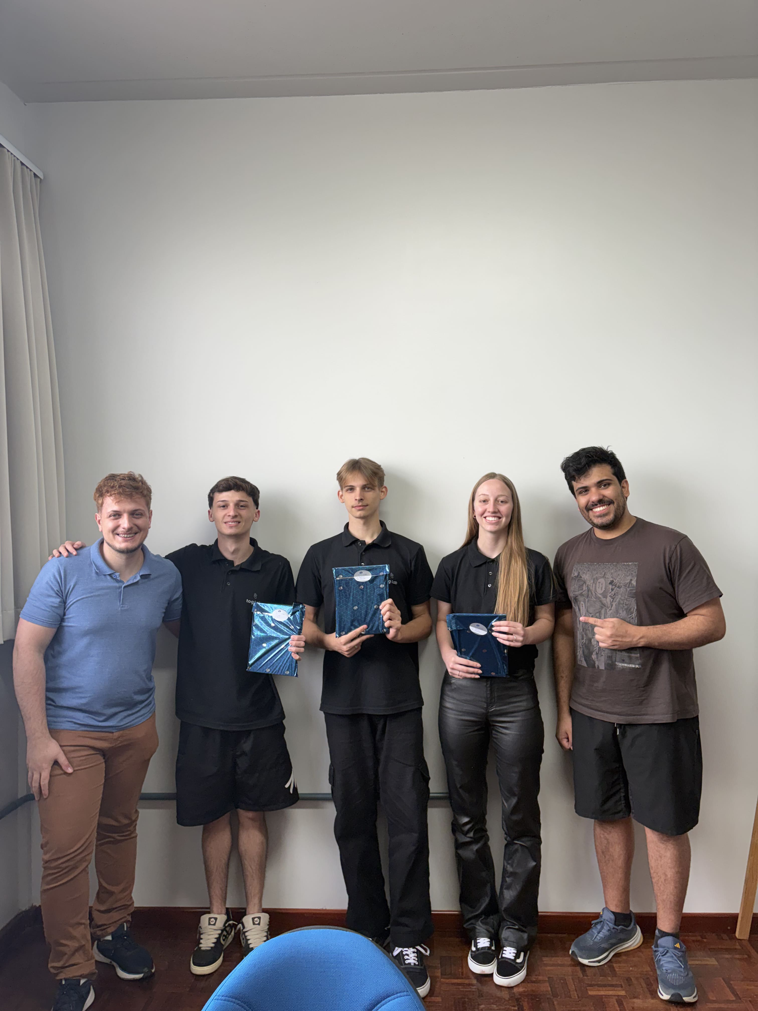 Cinco membros da equipe lado a lado em ambiente interno, alguns segurando cadernos azuis personalizados e sorrindo para a câmera