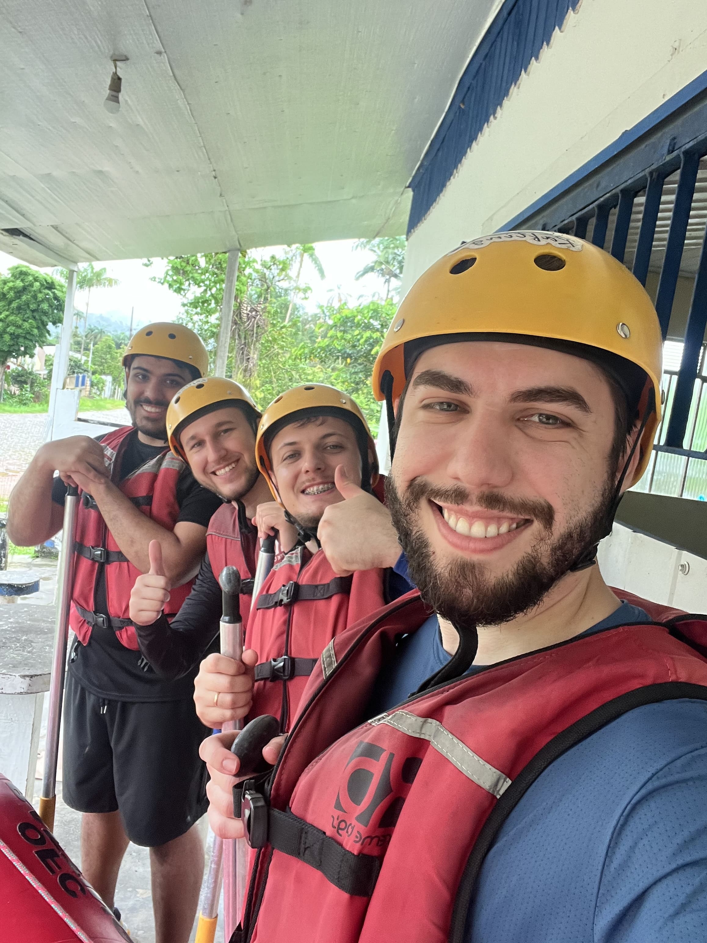 Quatro membros da equipe usando capacetes e coletes salva-vidas sorrindo e fazendo joinha enquanto seguram remos antes de uma atividade de rafting.