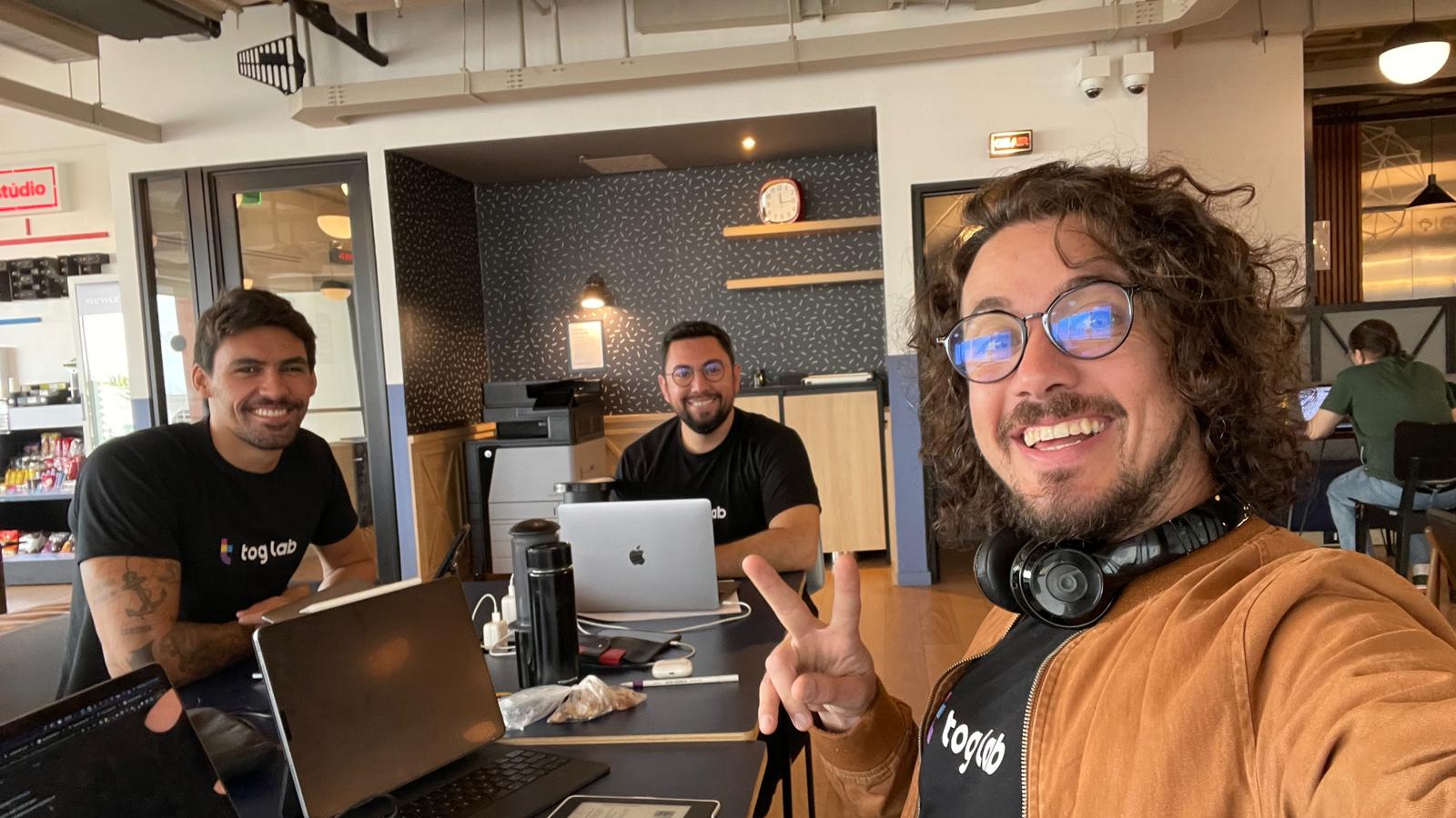 Tog Lab team working and smiling in an office environment. Three men smiling and working on laptops in an office environment. Two are seated at the table, and one is taking a selfie. All are wearing tog lab logo t-shirts.