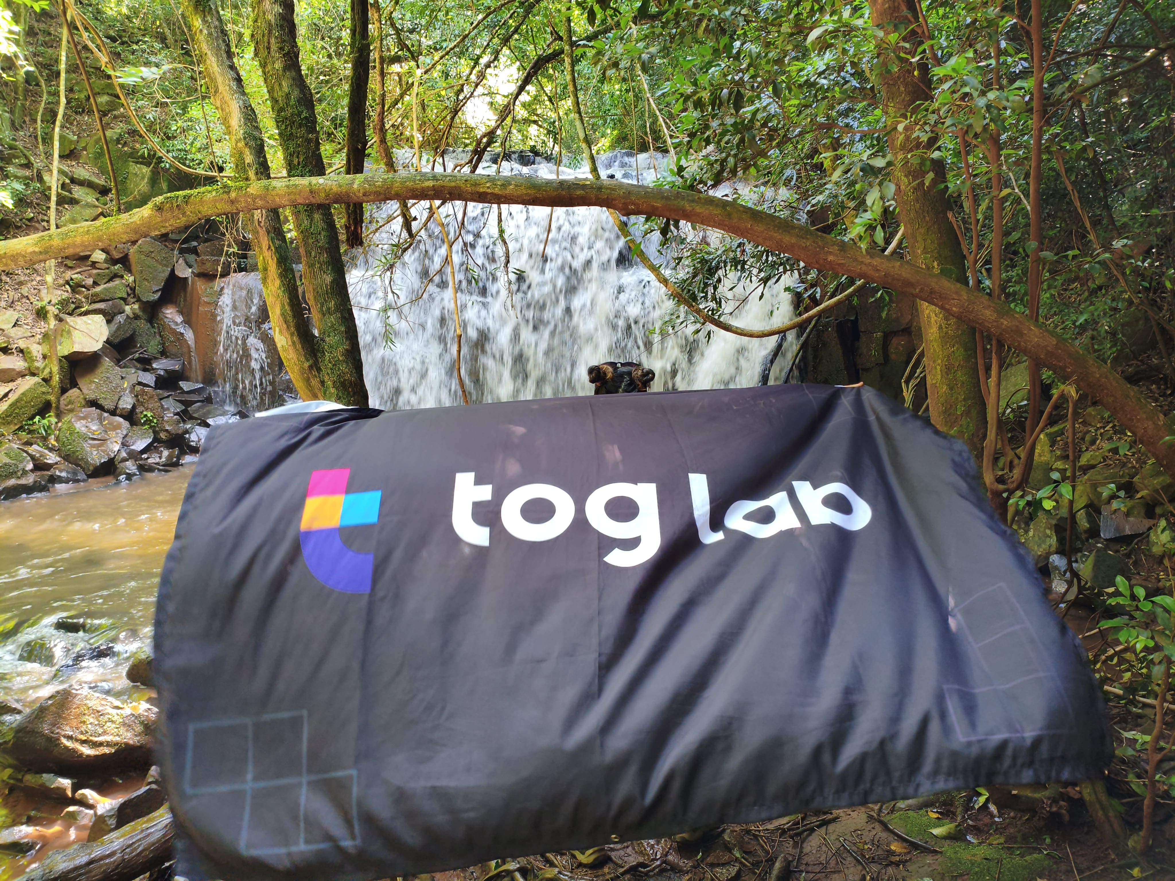 Uma bandeira da Toglab está estendida em frente a uma cachoeira, cercada por vegetação em meio à natureza.