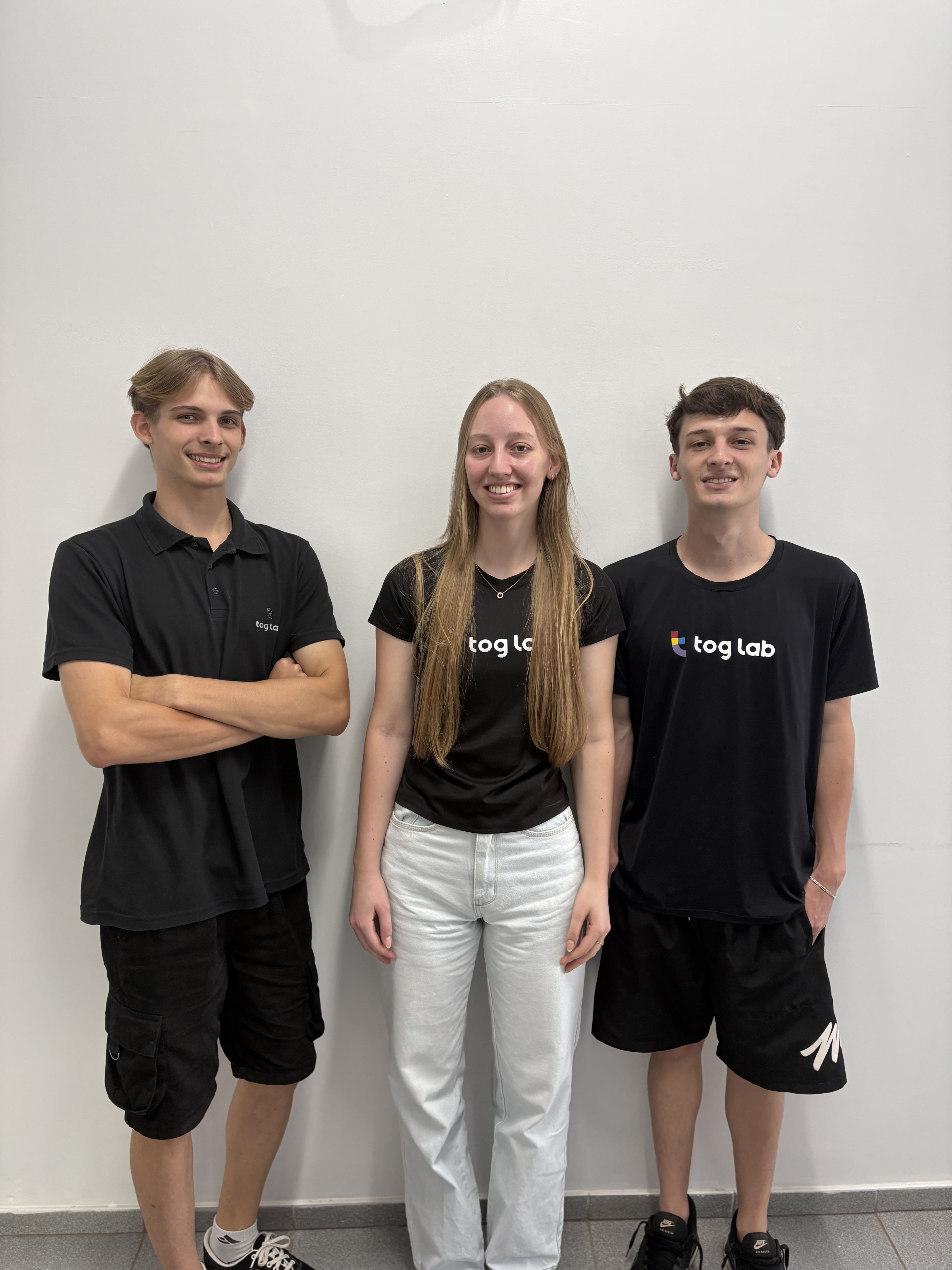 Três membros da equipe da Tog Lab lado a lado usando camisetas pretas da empresa contra uma parede clara.